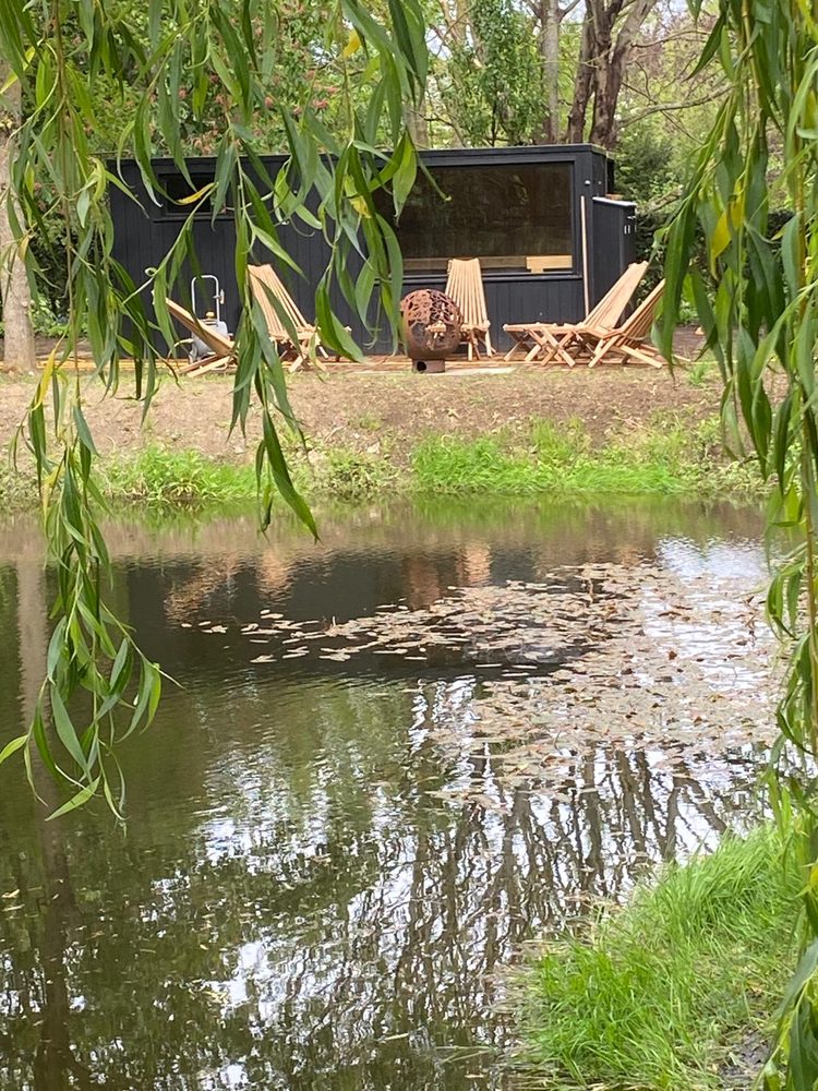 A Scandinavian-style black-clad lakeside sauna among mature willow in a Suffolk garden